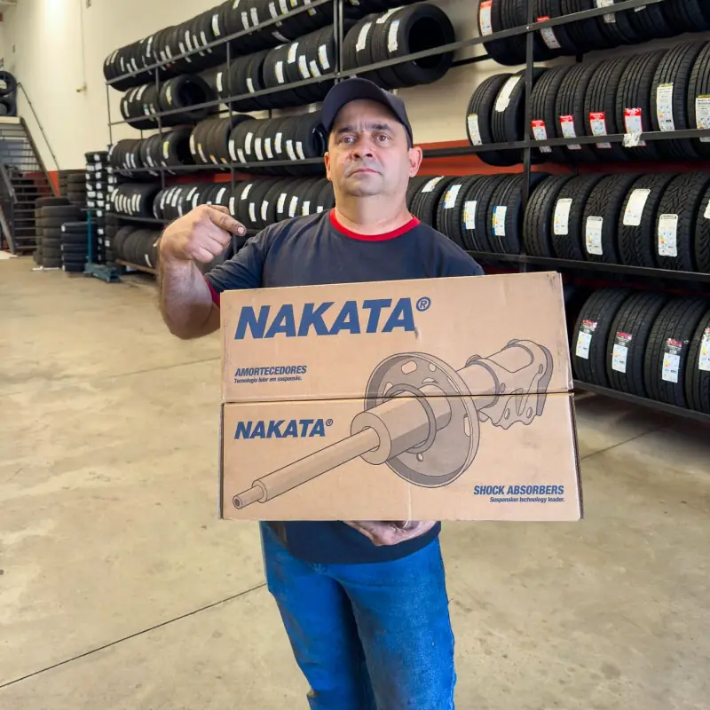 Mecânico do Jura Auto Center segurando caixas de amortecedores, com a parede de pneus ao fundo